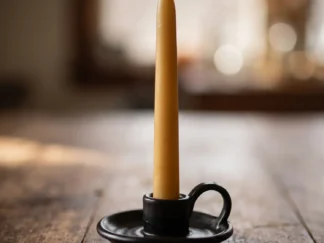 Pure beeswax taper candle in natural golden yellow, standing in a black chamberstick candle holder on a wooden table